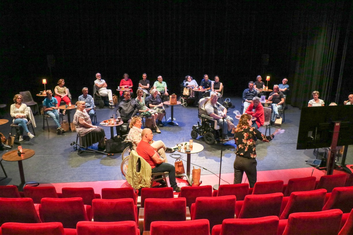Op het podium van de kleine zaal zitten ongeveer 30 mensen op anderhalve meter van elkaar.